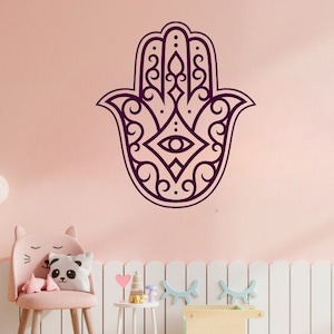 Può includere: Un adesivo da parete a forma di mano di Hamsa di colore viola con dettagli intricati e un occhio al centro. L'adesivo è su una parete rosa.