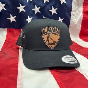 May include: Black and brown baseball cap with a patch that says "Lawn Enforcement Officer" and a silhouette of a person mowing a lawn. The cap has a mesh back.