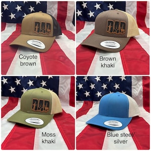 May include: Four trucker hats with a brown leather patch that says "DAD" in a stylized font. The hats are in four different colors: coyote brown, brown khaki, moss khaki, and blue steel/silver.