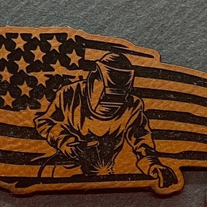 May include: A brown leather patch with a black silhouette of a welder wearing a welding mask and welding on a piece of metal. The background is a distressed American flag.