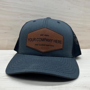 May include: A gray trucker hat with a black mesh back and a curved brim. The hat features a brown leather patch with the text "YOUR COMPANY HERE" and "EST. 2001". The bottom of the patch reads "Your custom text here".