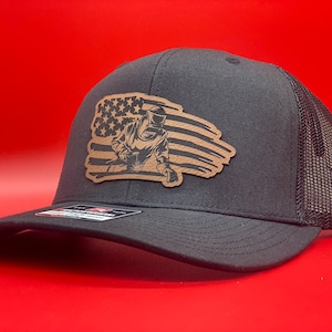 May include: A black and brown trucker hat with a distressed American flag design featuring a welder. The hat has a mesh back.