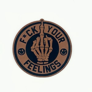 May include: A round brown sticker with a black border and the text "F*CK YOUR FEELINGS" in a black circle. A black skeleton hand with the middle finger extended is in the center of the circle.