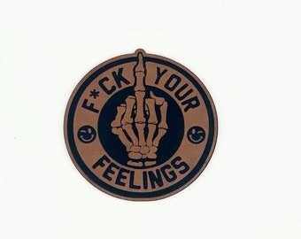 May include: A round brown sticker with a black border and the text "F*CK YOUR FEELINGS" in a black circle. A black skeleton hand with the middle finger extended is in the center of the circle.