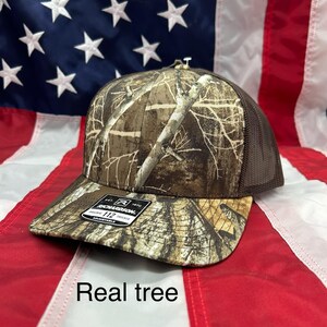 May include: A brown and tan camouflage trucker hat with a brown mesh back. The hat has a white label with the text "Richardson 112" on it. The hat is on a red, white, and blue American flag background. The text "Real tree" is written below the hat.