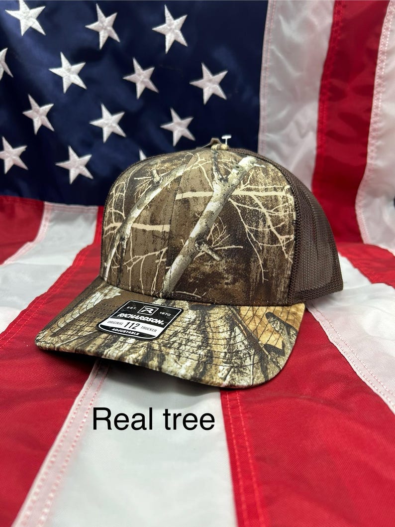May include: A brown and tan camouflage trucker hat with a brown mesh back. The hat has a white label with the text "Richardson 112" on it. The hat is on a red, white, and blue American flag background. The text "Real tree" is written below the hat.