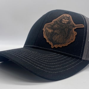 May include: Black and gray trucker hat with a brown leather patch featuring a skeleton figure holding a rifle. The patch is in the style of a grim reaper.