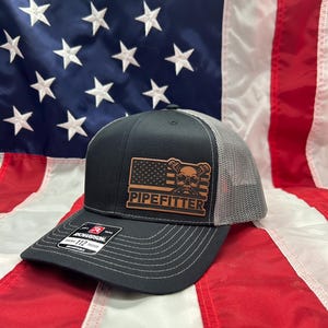 May include: A black and grey trucker hat with a leather patch that says "Pipefitter" and a skull and crossbones design. The patch is on a red, white, and blue American flag background.
