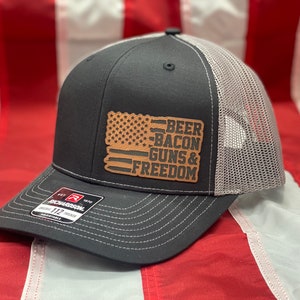 May include: A black and gray trucker hat with a brown leather patch featuring an American flag and the text "BEER BACON GUNS & FREEDOM".