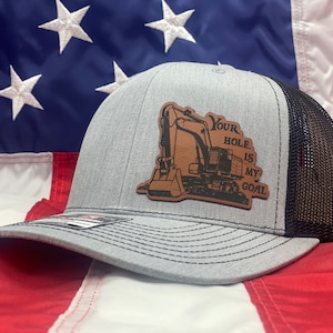 May include: A gray and black trucker hat with a brown leather patch featuring an excavator and the text "Your Hole Is My Goal".
