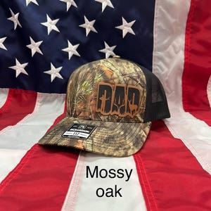 May include: A camouflage baseball cap with a brown leather patch that says "DAD" with a cross and a deer silhouette. The cap is on a red, white, and blue American flag. The cap is labeled "Mossy Oak".