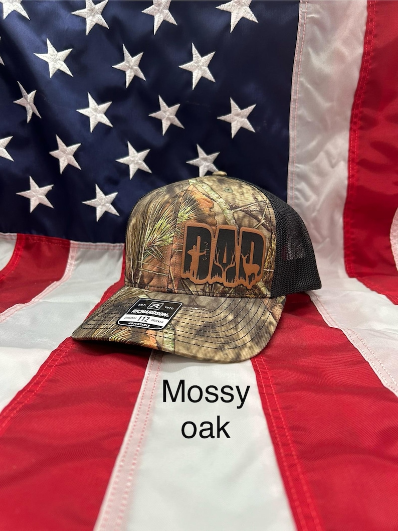 May include: A camouflage baseball cap with a brown leather patch that says "DAD" with a cross and a deer silhouette. The cap is on a red, white, and blue American flag. The cap is labeled "Mossy Oak".