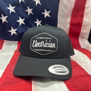 May include: Black snapback hat with a mesh back and a curved brim. The front of the hat features a white graphic that reads "BADASS Electrician BLUECOLLAR". A white tag with "SNAPBACK" is on the brim.