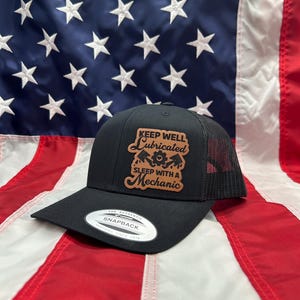 May include: Black snapback trucker hat with a mesh back and a curved brim. The hat features a brown leather patch with the text "KEEP WELL LUBRICATED SLEEP WITH A MECHANIC" and mechanic tools. The hat is set against an American flag.
