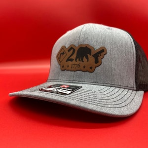 May include: A gray and black trucker hat with a brown leather patch featuring a bear, a gun, and the text "2A 1776".