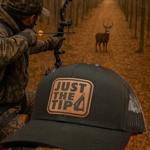 May include: Black trucker hat with a leather patch that reads "JUST THE TIP" in white and brown. The hat is in the foreground, with a person aiming a bow and arrow at a deer in the background. The scene is set in a forest.