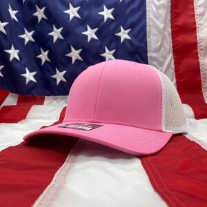 May include: A pink and white trucker hat with a mesh back. The hat is lying on an American flag.