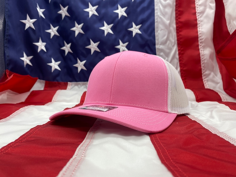 May include: A pink and white trucker hat with a mesh back. The hat is lying on an American flag.