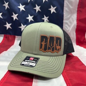 May include: A green and black trucker hat with a brown leather patch that says "DAD" with a silhouette of a hunter and deer. The hat is made by Richardson 112.