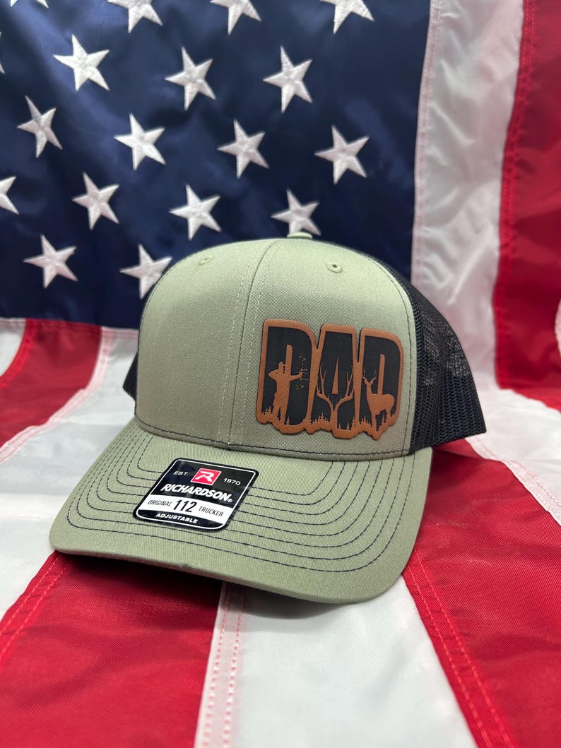 May include: A green and black trucker hat with a brown leather patch that says "DAD" with a silhouette of a hunter and deer. The hat is made by Richardson 112.