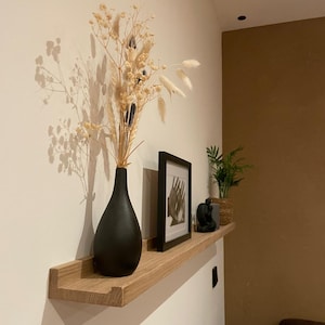 Könnte beinhalten: Ein Holzregal mit einer schwarzen Vase gefüllt mit Trockenblumen, einem schwarzen gerahmten Bild und einem kleinen Dekorationsobjekt. Das Regal ist an einer weißen Wand montiert.