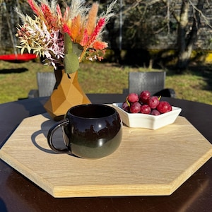 Könnte beinhalten: Eine dunkelbraune Keramiktasse steht auf einem hellbraunen Holztablett mit einer weißen Schale voller roter Trauben. Das Tablett befindet sich auf einem dunkelbraunen Tisch.