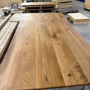 May include: A large, rectangular wooden table with a natural wood grain finish. The table is made of light brown wood and has a smooth surface. The table is supported by metal legs. Other wooden items are in the background.