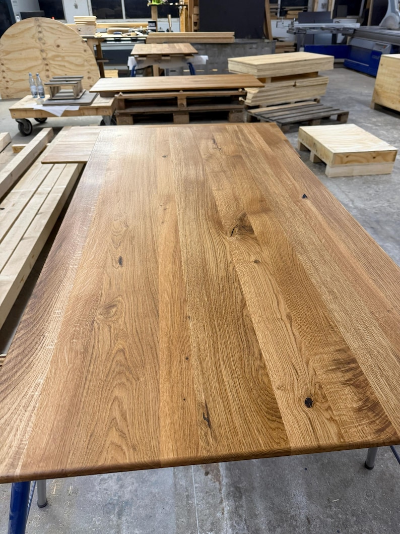 May include: A large, rectangular wooden table with a natural wood grain finish. The table is made of light brown wood and has a smooth surface. The table is supported by metal legs. Other wooden items are in the background.