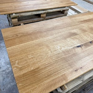 May include: Two unfinished, light-brown wooden table tops. The wood grain is visible, and the tables are rectangular. One table is on top of a wooden pallet, and the other is in the foreground.
