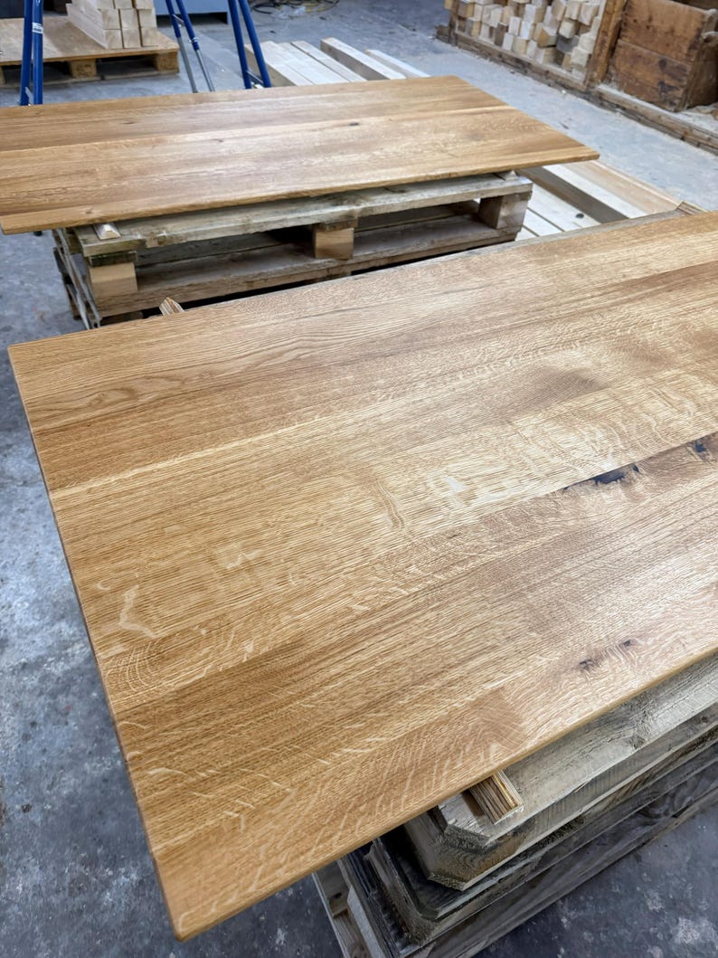 May include: Two unfinished, light-brown wooden table tops. The wood grain is visible, and the tables are rectangular. One table is on top of a wooden pallet, and the other is in the foreground.