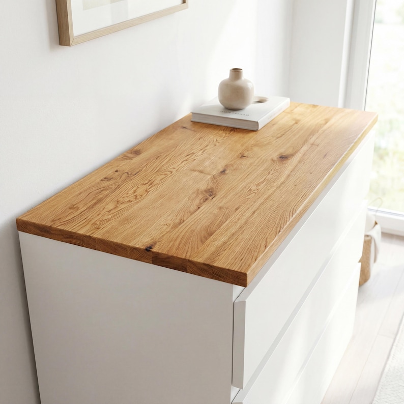 May include: A white dresser with three drawers and a light brown wooden top. A small beige vase and a book are on top of the dresser. The dresser is in front of a white wall.