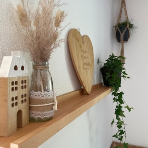 Op de afbeelding: Een houten plank met een hartvormig snijplank, een glazen vaas met gedroogd pampasgras, een klein houten huisje en een hangplant. De snijplank heeft de tekst "Gl&uuml;ckliche Herzen, beste Zeit f&uuml;r Familie" erop gegraveerd.