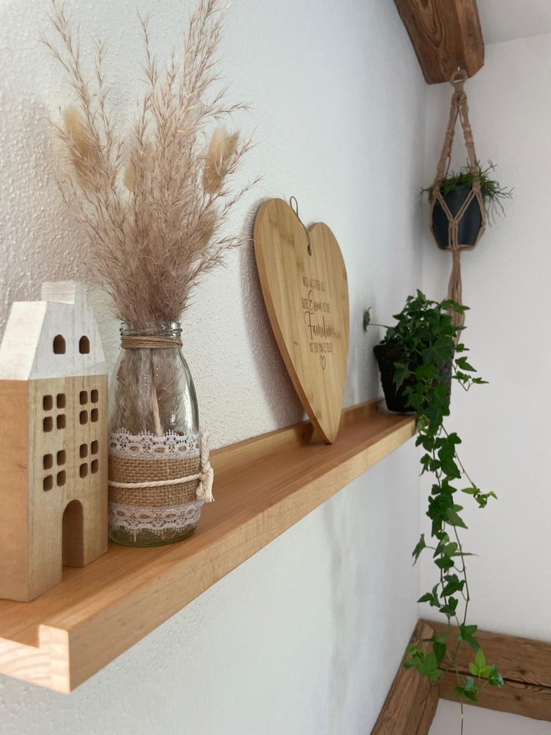 Op de afbeelding: Een houten plank met een hartvormig snijplank, een glazen vaas met gedroogd pampasgras, een klein houten huisje en een hangplant. De snijplank heeft de tekst "Gl&uuml;ckliche Herzen, beste Zeit f&uuml;r Familie" erop gegraveerd.