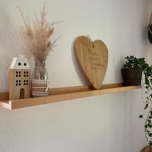 Op de afbeelding: Een houten plank met een hartvormig snijplank waarop "Welkom in onze familie" staat, een klein huisvormig beeldje, een vaas met gedroogde bloemen en een hangende potplant.