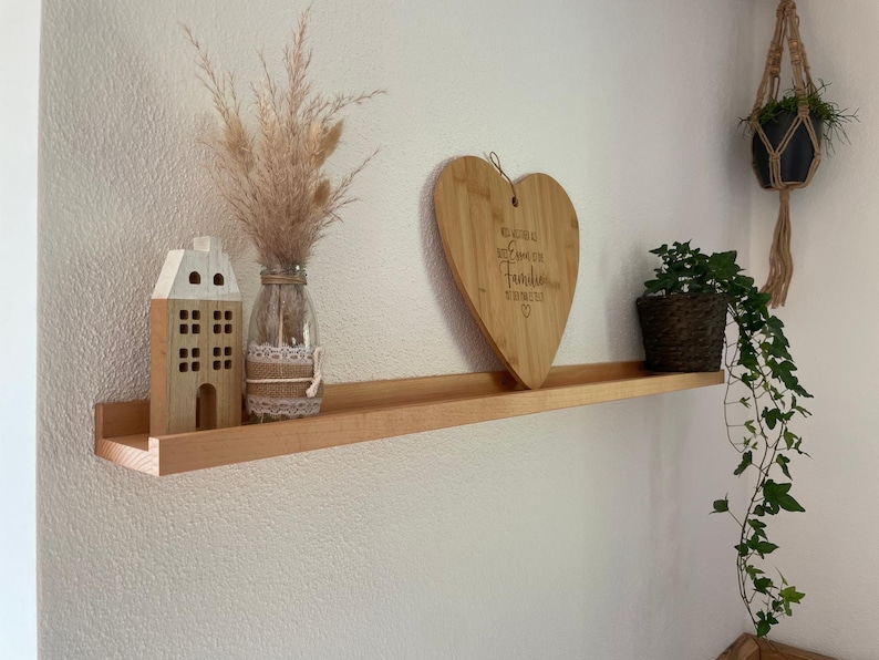 Op de afbeelding: Een houten plank met een hartvormig snijplank waarop "Welkom in onze familie" staat, een klein huisvormig beeldje, een vaas met gedroogde bloemen en een hangende potplant.