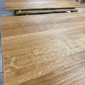 May include: Close-up of two unfinished wooden table tops. The wood grain is visible, with a light brown color and natural variations. The tables are rectangular and appear to be ready for finishing or assembly. The wood has a natural, rustic appearance.