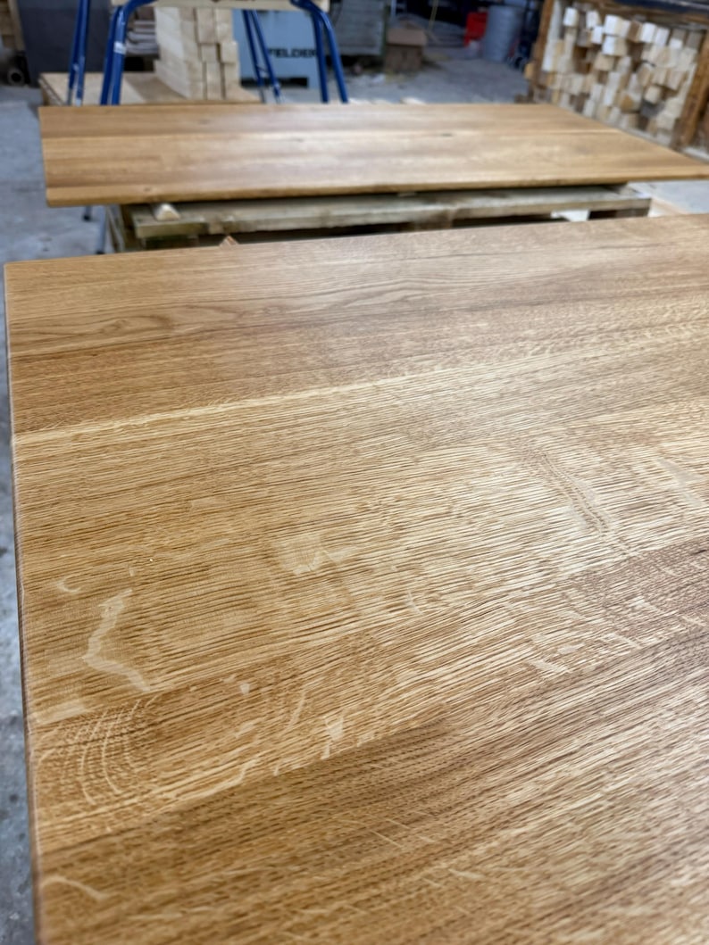 May include: Close-up of two unfinished wooden table tops. The wood grain is visible, with a light brown color and natural variations. The tables are rectangular and appear to be ready for finishing or assembly. The wood has a natural, rustic appearance.