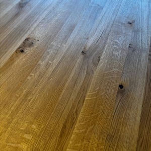 May include: Close-up of a wooden surface with a natural wood grain pattern. The wood has a warm, golden-brown hue with darker knots and variations in color. The planks are arranged in parallel lines, creating a textured and visually appealing surface.