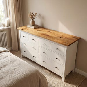 May include: A white dresser with eight drawers and a natural wood top. The dresser has black knobs and is in a bedroom setting. A vase with dried flowers sits on top of the dresser.