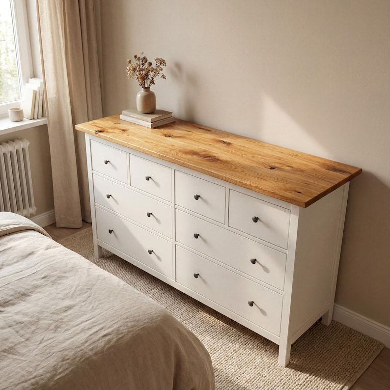 May include: A white dresser with eight drawers and a natural wood top. The dresser has black knobs and is in a bedroom setting. A vase with dried flowers sits on top of the dresser.