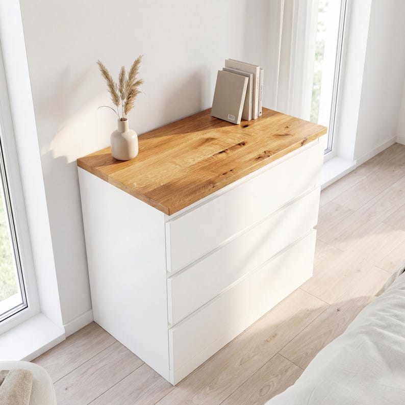May include: A white three-drawer dresser with a natural wood top. The dresser has a minimalist design, with clean lines and a simple aesthetic. A vase with dried flowers and a stack of books sit on top.