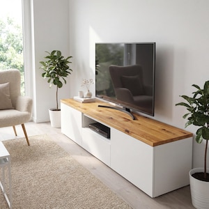 May include: A modern white TV stand with a natural wood top. The stand features two doors and an open shelf for media components. A large flat-screen TV sits on top. The room includes a beige rug and potted plants.