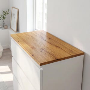 May include: A white dresser with a light brown wooden top. The wood grain is visible on the top surface. The dresser has four drawers and is positioned near a window.