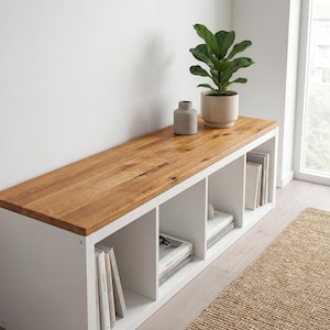 May include: A white storage bench with a natural wood top. The bench has four cubby spaces filled with books and decorative items. A potted plant and a small vase sit on top of the bench.