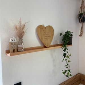 Op de afbeelding: Een houten plank met een hartvormig snijplank waarop "Wir wachsen alle mit einem St&uuml;ck Familie" staat. Op de plank staan een klein wit huis, een vaas met gedroogde bloemen en een potplant.
