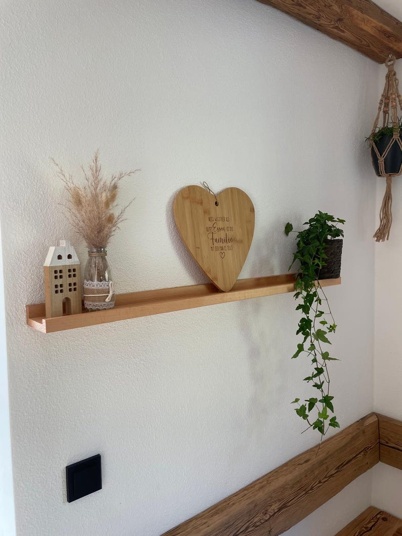 Op de afbeelding: Een houten plank met een hartvormig snijplank waarop "Wir wachsen alle mit einem St&uuml;ck Familie" staat. Op de plank staan een klein wit huis, een vaas met gedroogde bloemen en een potplant.