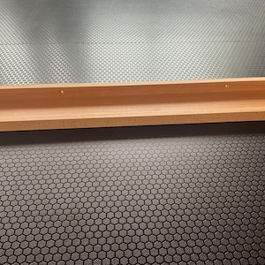 Op de afbeelding: Een houten plank met een lichte bocht aan de voorkant. De plank is lichtbruin en heeft twee kleine gaatjes aan de bovenkant.
