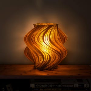 Puede incluir: Una lámpara decorativa con un diseño único y arremolinado. La lámpara está hecha de un material cálido de color dorado, con una apariencia texturizada y en capas. La luz brilla a través de las capas, creando un brillo suave y acogedor. La lámpara está sobre una estantería de madera.