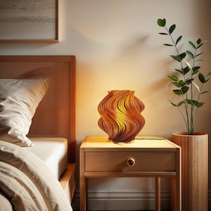 May include: A table lamp with a spiraling, wave-like structure featuring a gradient of warm orange, yellow, and brown tones. The lamp emits a soft, glowing light from within, casting a warm illumination across the surface it rests upon.