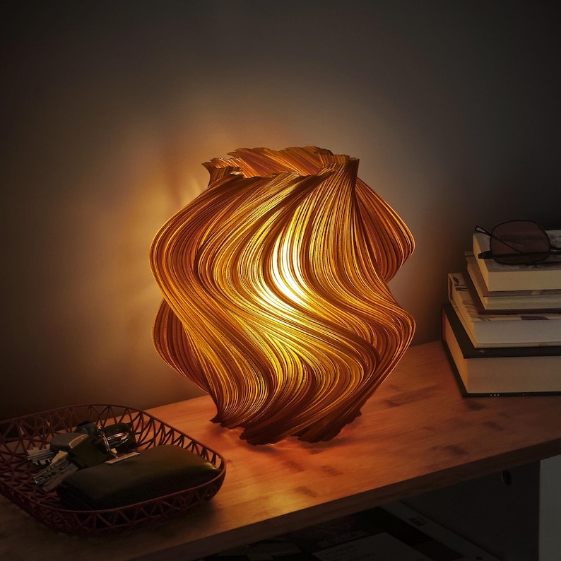 May include: A table lamp with a warm, glowing light emanating from within a textured, amber-colored shade featuring vertical, undulating, wave-like ridges that create a dynamic, spiraling pattern across the entire surface of the lamp.
